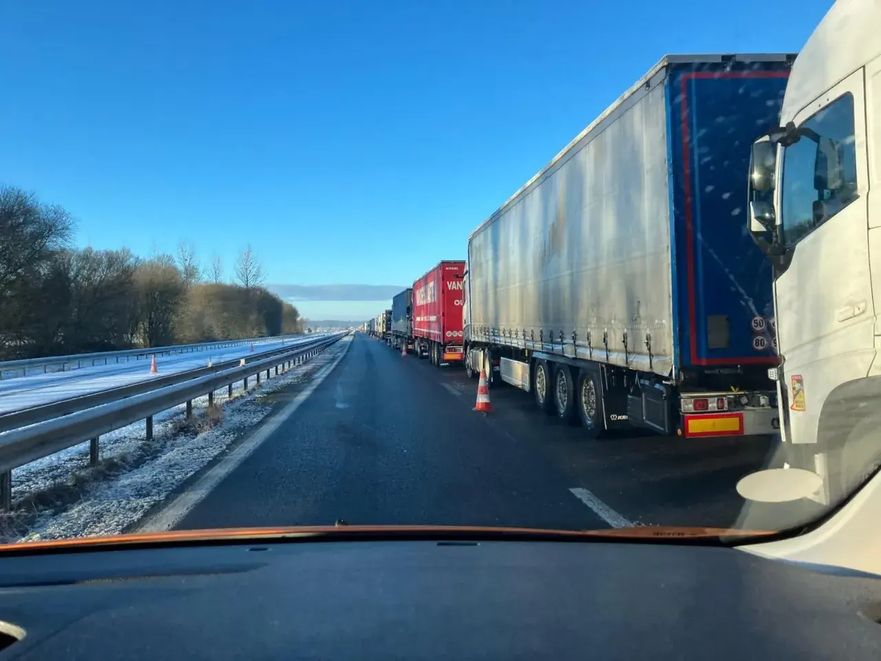 Longue file de véhicules poids lourds à l'arrêt sur une autoroute enneigée sous un ciel bleu clair.
