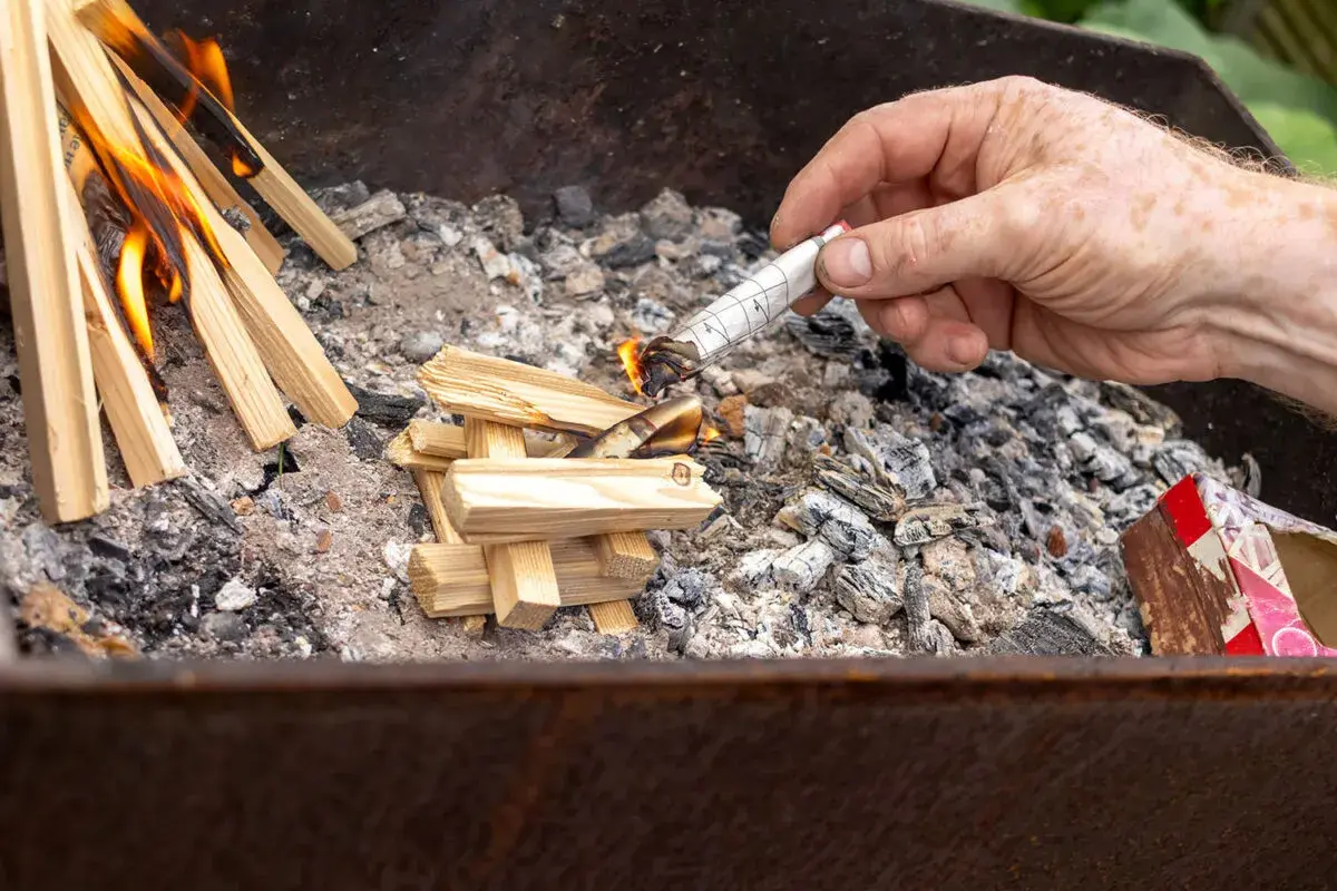 Maintenant, comment allumer un barbecue au charbon : une main allume un morceau de papier roulé pour enflammer des brindilles dans le brasero.