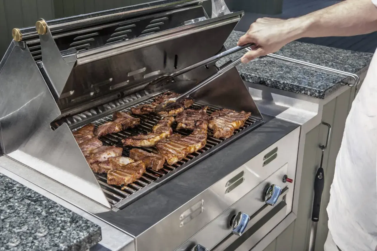 Un homme utilise une pince pour retourner des côtes levées sur un gril à gaz. L'odeur alléchante de la viande cuite emplit l'air.