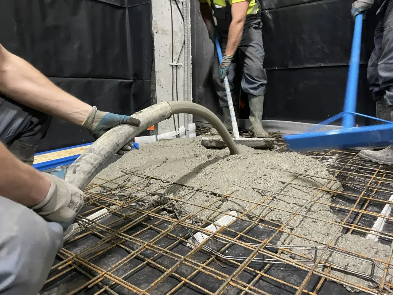 Pracownicy wykonują pompowanie betonu na zbrojenie. Beton płynie z węża, a drugi pracownik rozprowadza go łopatą.