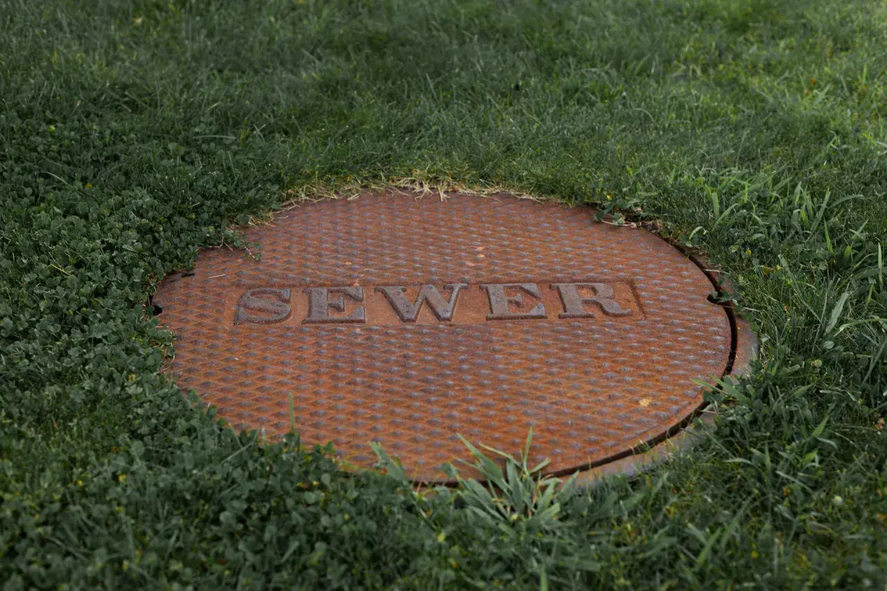 Zardzewiała pokrywa szamba z napisem "SEWER" w trawie. Czy na szambo trzeba mieć pozwolenie?