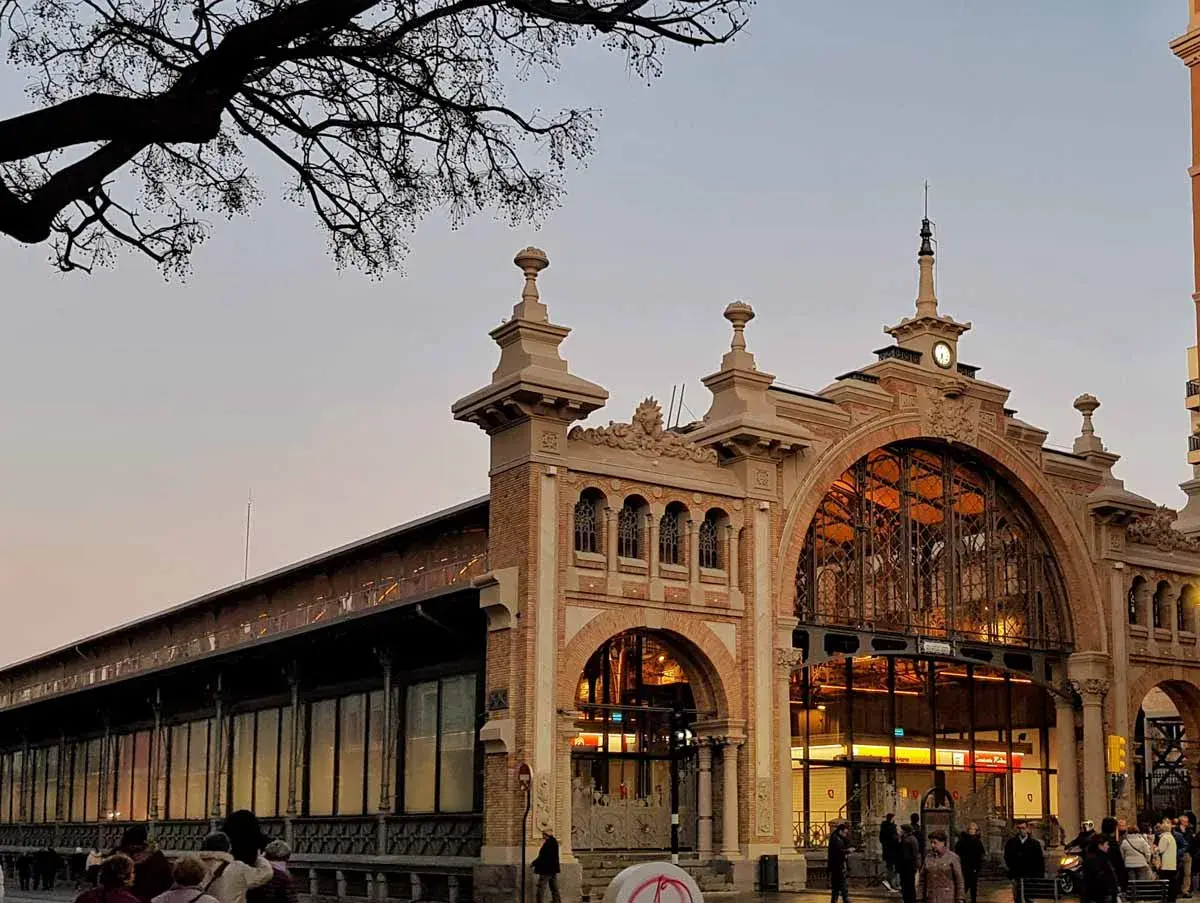 El **mercado central Zaragoza** con su arquitectura de hierro y cristal, iluminado al atardecer. Gente pasea por la entrada principal.