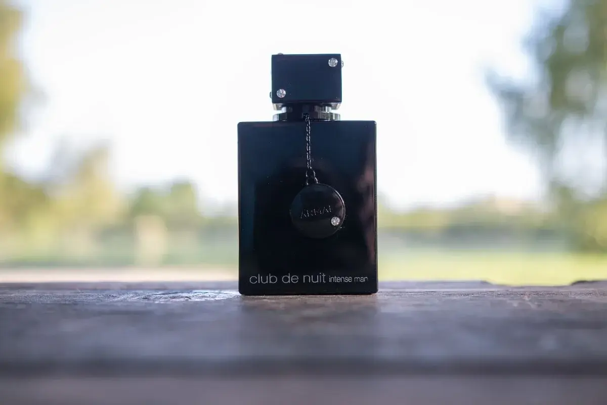 Elegancka czarna butelka perfum Club de Nuit Intense Man, ozdobiona łańcuszkiem i dyskiem z logo, stoi na drewnianym stole.