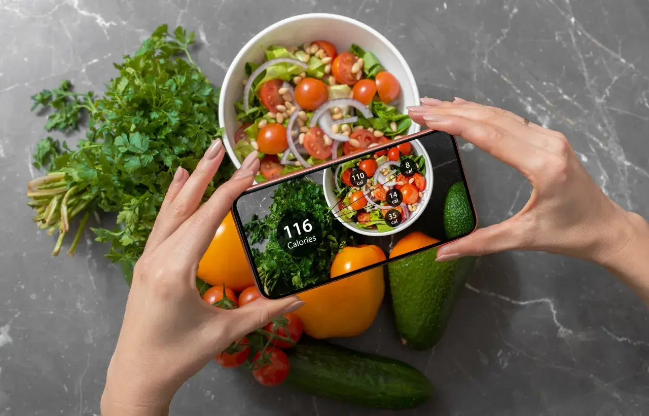 Ręce trzymają smartfon z aplikacją pokazującą kalorie. Na ekranie widać sałatkę i zioła, a obok na blacie leżą warzywa. Zastanawiasz się, ile kalorii żeby przytyć?