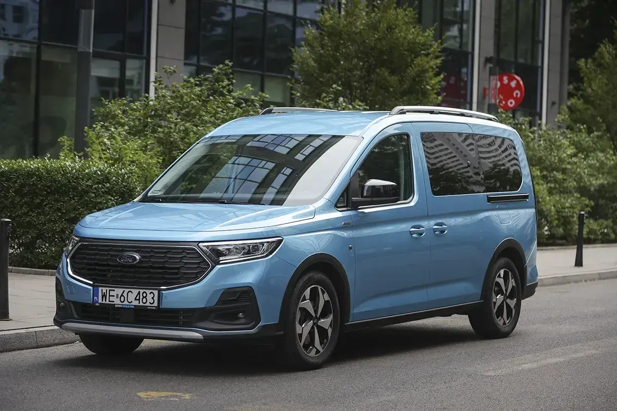 Błękitny ford 7 osobowy na tle nowoczesnego budynku. Idealny dla rodziny na wakacyjne wyprawy.