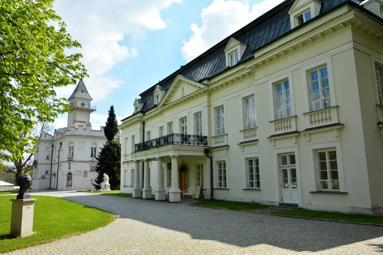 Pałac Radziejowice noclegi cennik: biały pałac z wieżą, zielony trawnik, rzeźby, drzewa, błękitne niebo z chmurami.