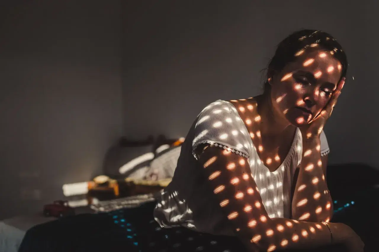 Vrouw met gesloten ogen, haar gezicht en armen bedekt met schaduwen van lichtplekken, alsof ze zich terugtrekt, niet tegen drukte kan.