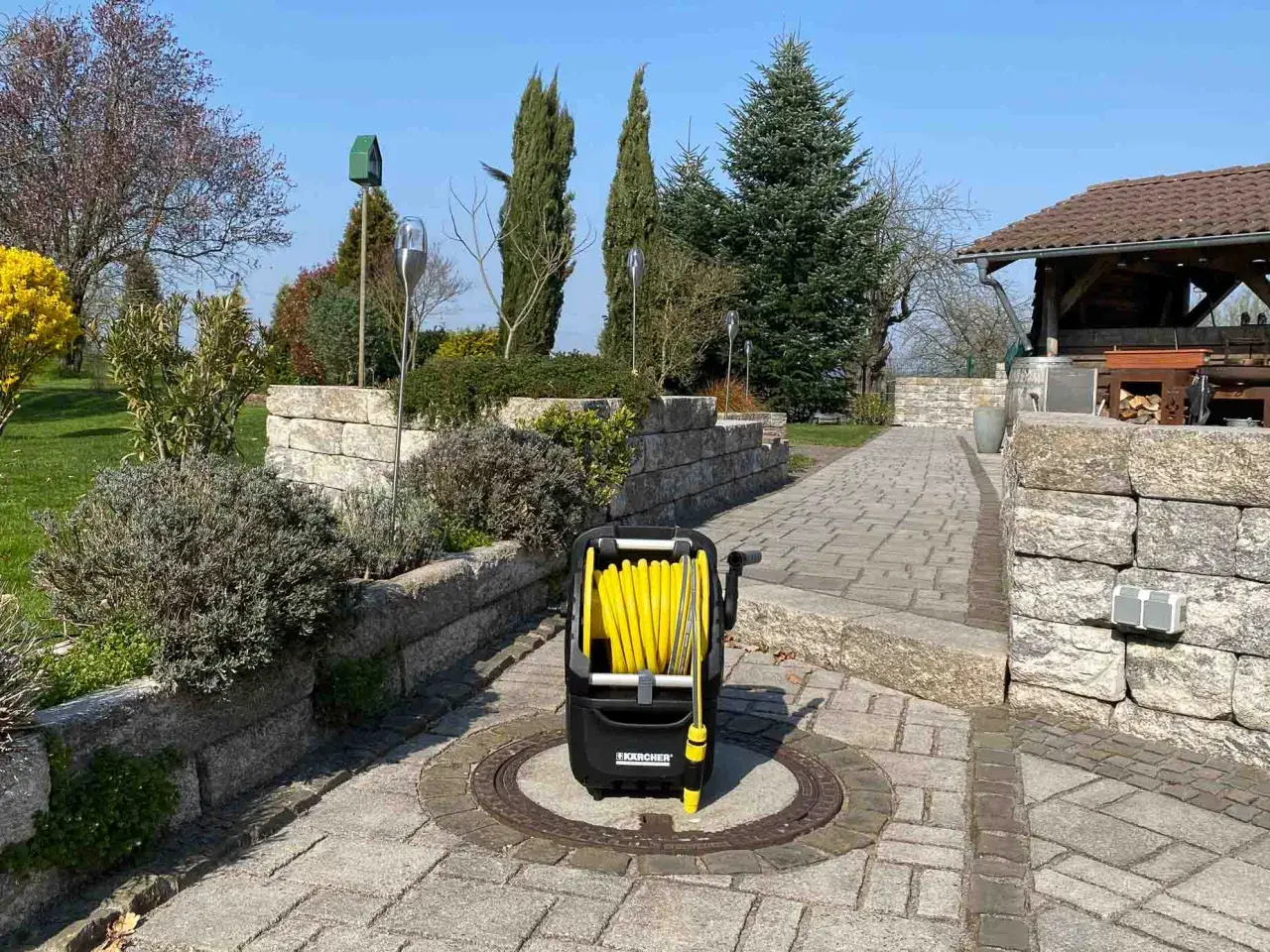 Ein gelber Schlauchwagen steht auf einem gepflasterten Weg im Garten, bereit für die Gartenarbeit.