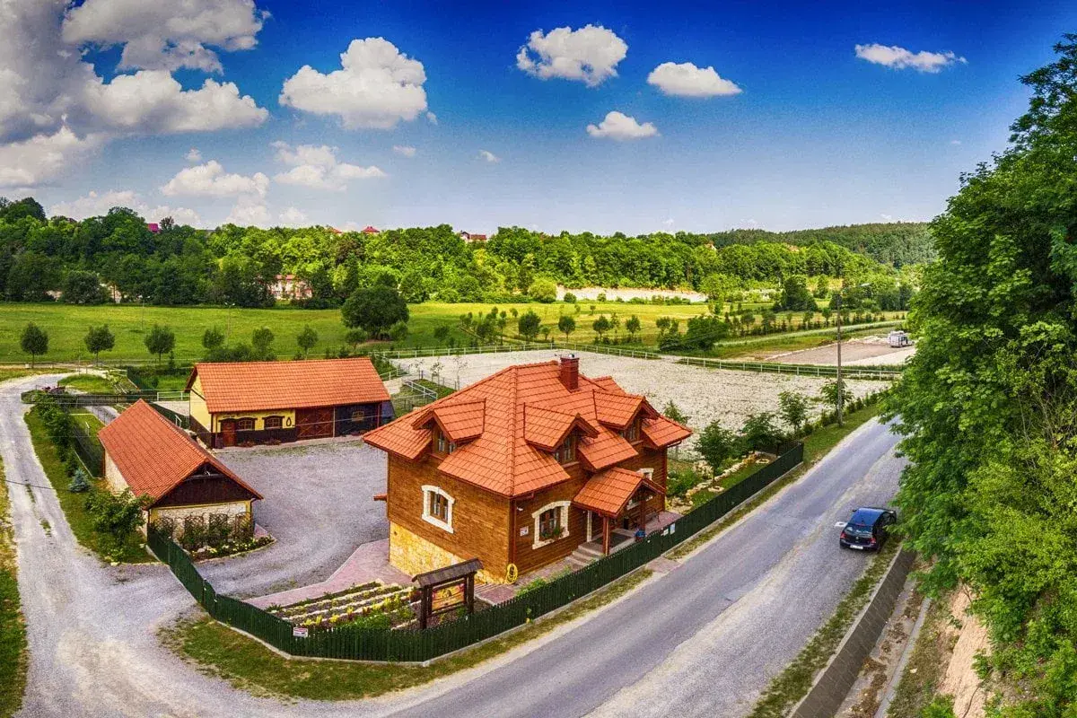 Drewniane budynki rancza z czerwonymi dachami, otoczone zielenią, pod błękitnym niebem z chmurami.