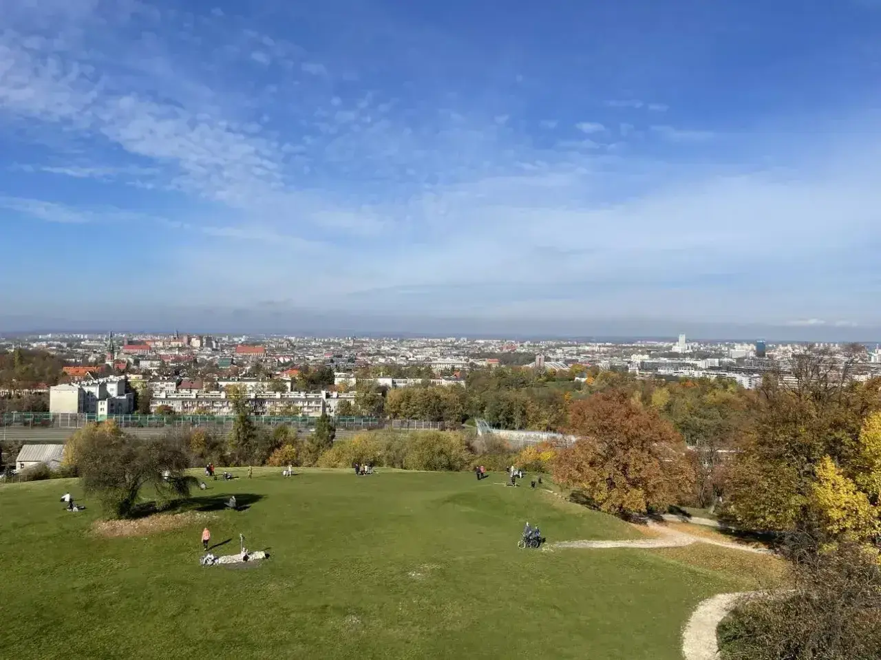 Widok na Kraków z zielonego wzgórza, gdzie ludzie odpoczywają. W oddali rozciąga się panorama miasta, a jesienne drzewa dodają uroku. To idealne miejsce na spacer, jak na **Kopcu Kraka**.