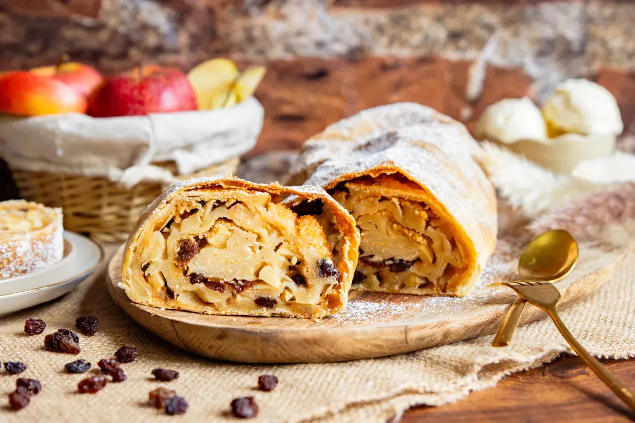 Ein köstlicher Apfelstrudel, wie ihn Landfrauen backen, mit saftiger Füllung und Puderzucker bestäubt. Dazu passen Rosinen und eine Kugel Eis.