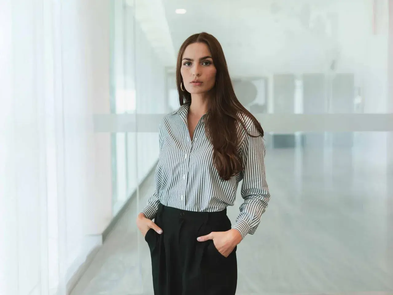 Frau in gestreifter Bluse und schwarzen Hosen, ein Look im business casual Stil, steht mit den Händen in den Taschen.