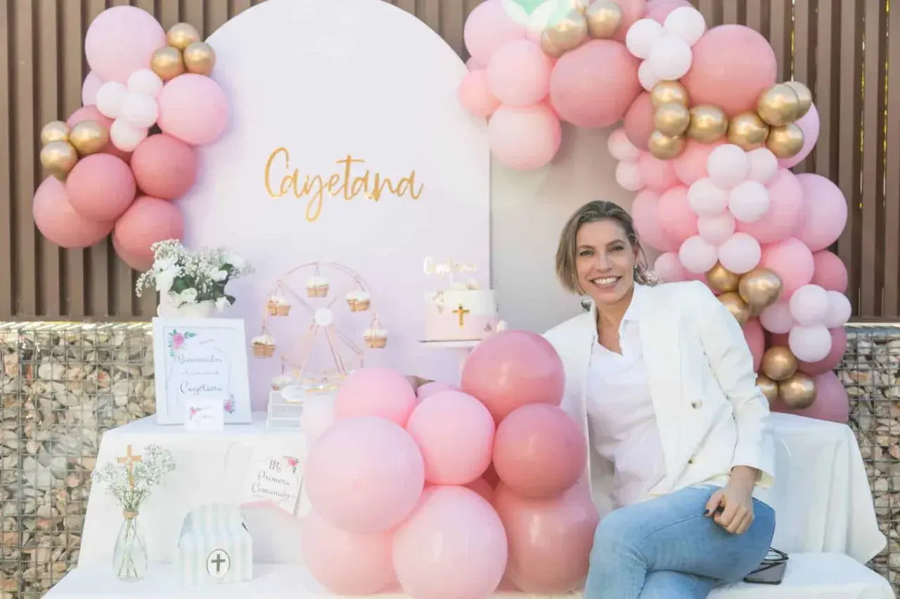 Una mujer sonríe junto a una mesa decorada para una comunión casera, con globos rosas y dorados, un pastel y un cartel de bienvenida.