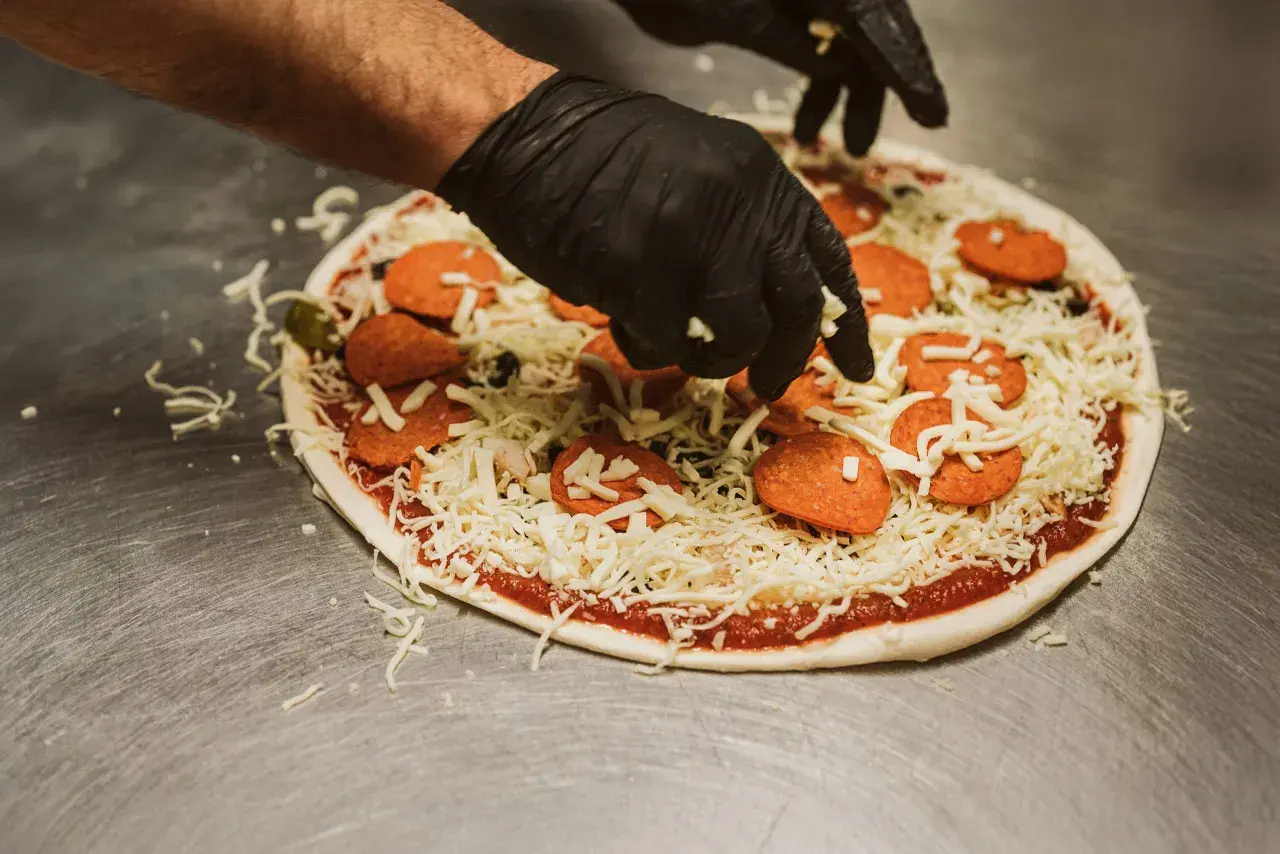Händer i svarta handskar lägger ost på en pizza med pepperoni och oliver. En klassisk kombination för härliga pizza smaker.