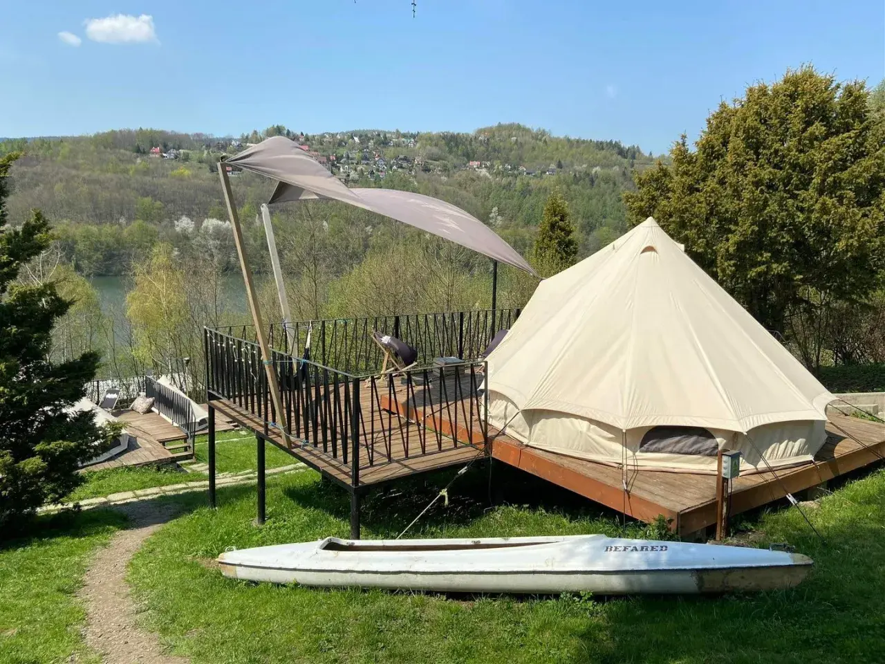 Namiot typu glamping camp z widokiem na jezioro i wzgórza. Na tarasie stolik i krzesło. Obok leży kajak.