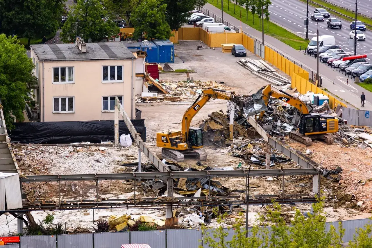 Dwa koparki pracują na placu budowy, rozbierając budynek. W tle widać drogę z samochodami i drzewa.