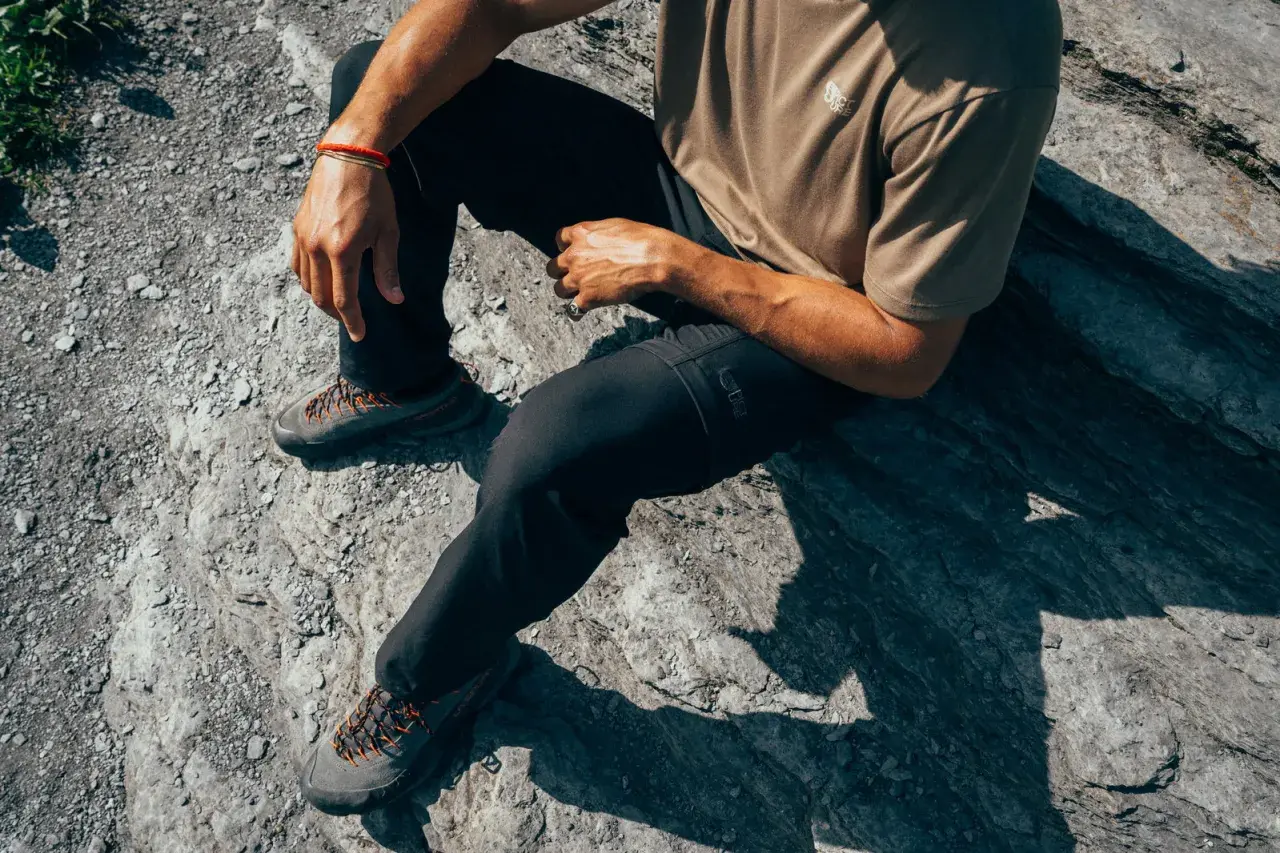 Man sitting on rocks, wearing black pants and hiking shoes. The shoes are grey with orange laces, perfect for schoenen oprekken on any terrain.