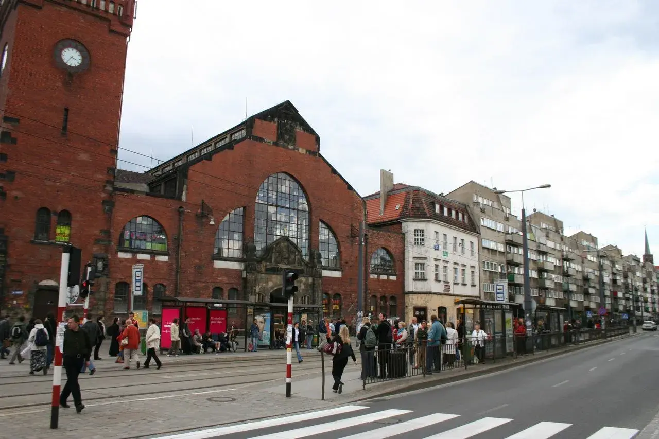 Menschen warten an einer Straßenbahnhaltestelle vor der imposanten Markthalle Breslau.