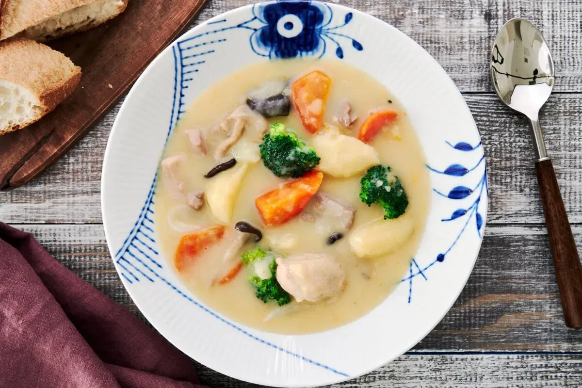 A hearty bowl of Japanese cream stew, brimming with chicken, carrots, potatoes, broccoli, and mushrooms, served with crusty bread.