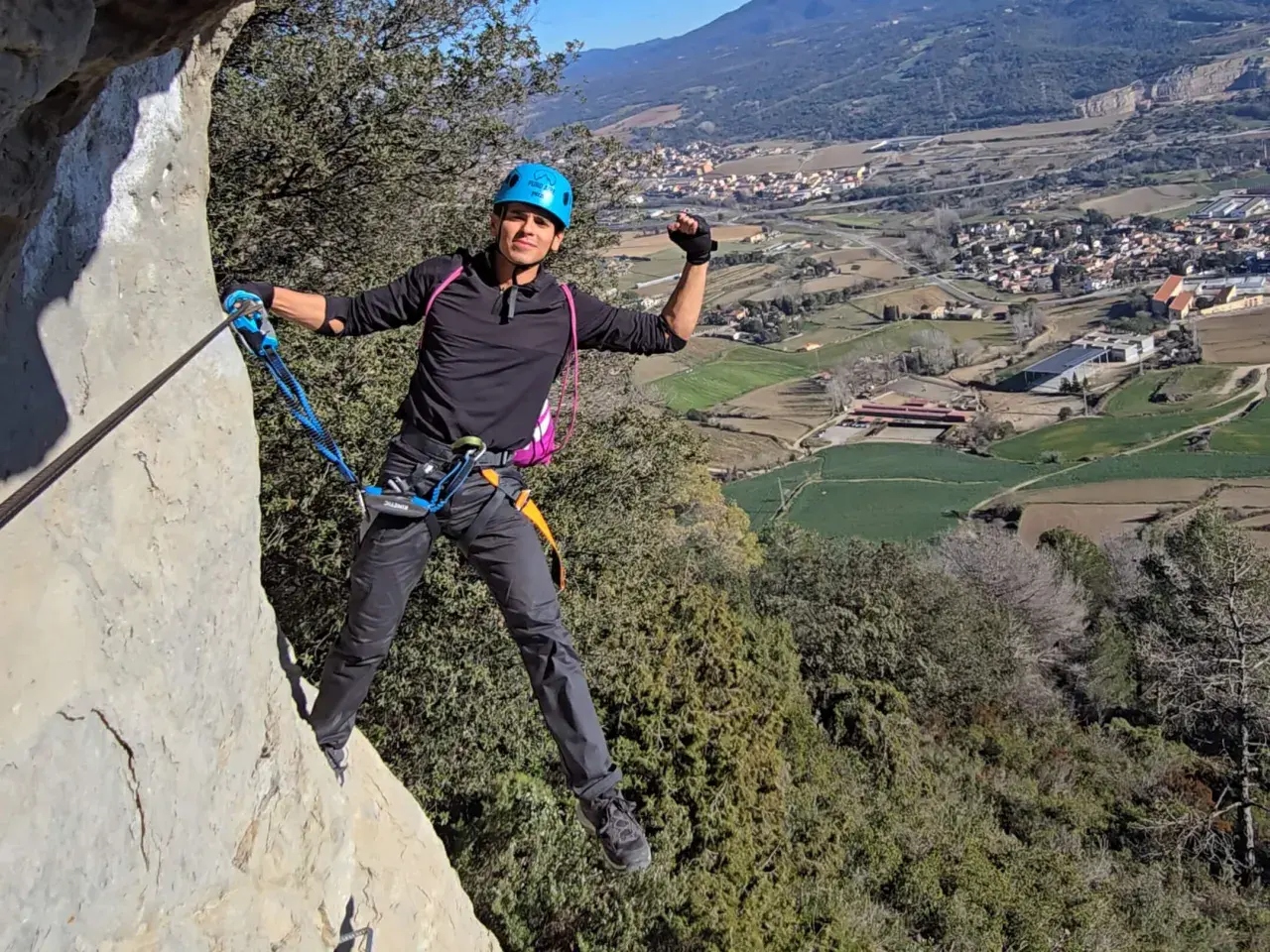Hombre con casco azul y arnés, asegurado a un cable de **material vía ferrata**, disfruta de las vistas del valle.