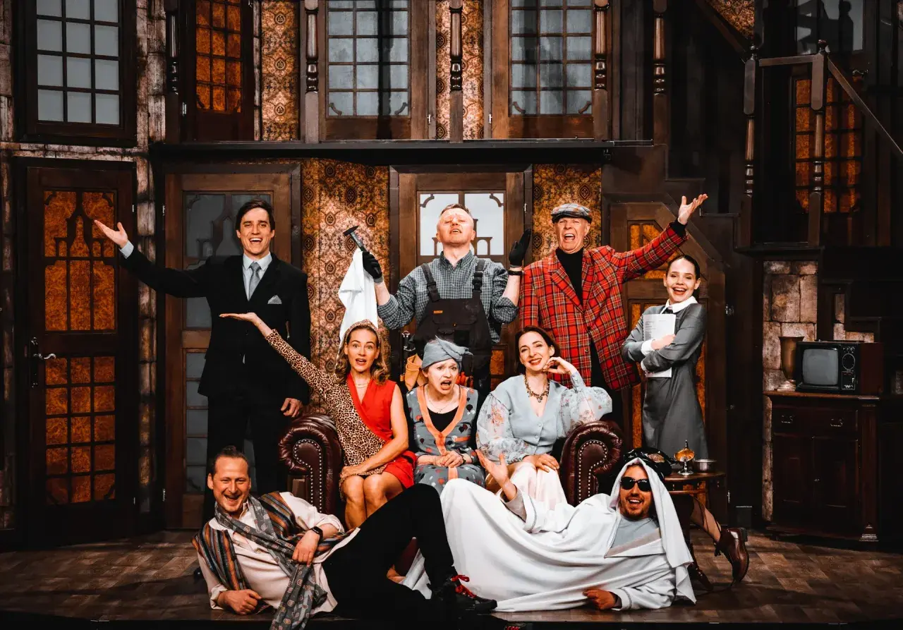 Aktorzy w teatralnej scenografii, jakby w teatrze bez rzędów, pozują do zdjęcia. Wszyscy uśmiechnięci, w barwnych strojach, tworzą radosną atmosferę.