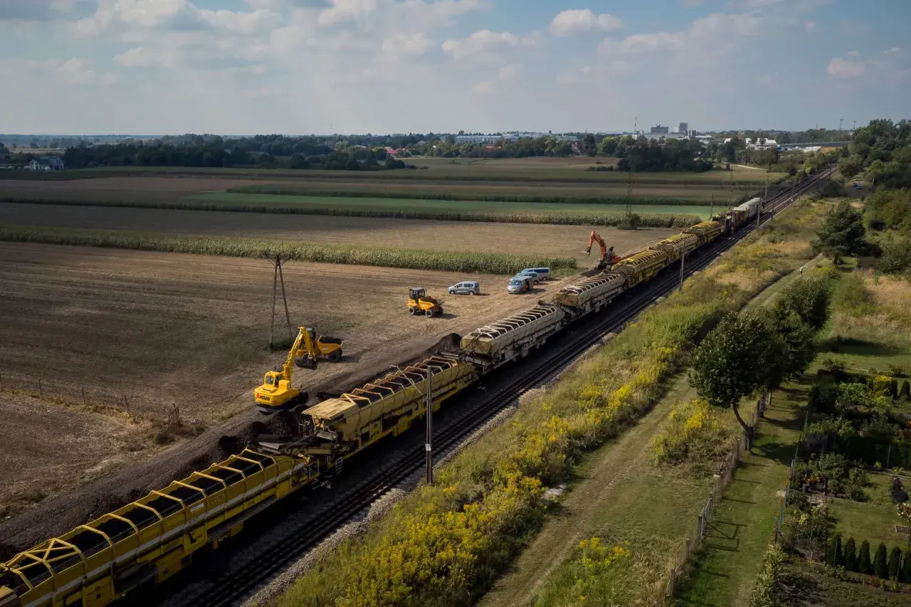 Długi pociąg z wagonami wypełnionymi ziemią, obok koparki i samochodów. Prace torowe w tle pól i zabudowań, widać odbojnica kolejowa.