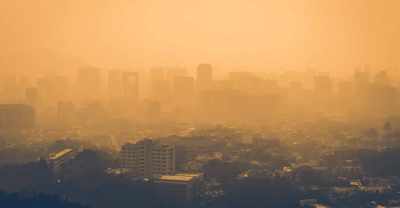 Vista aérea de una ciudad envuelta en una densa neblina anaranjada, con siluetas de edificios apenas visibles. Las partículas PM10 flotan en el aire, creando esta atmósfera opresiva.