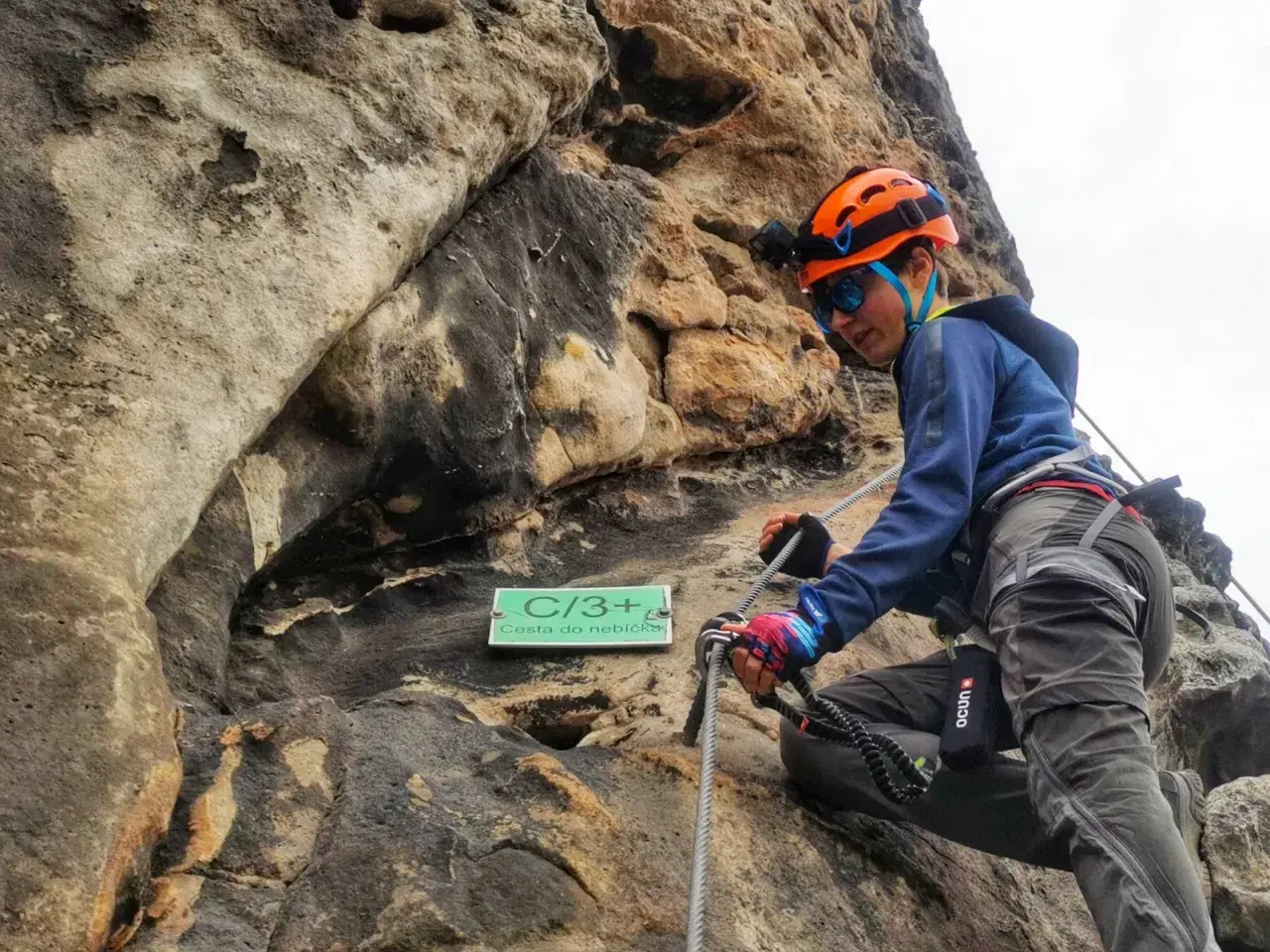 Młoda osoba w kasku wspina się po skale na via ferrata w Decinie. Tabliczka "Cesta do nebíčka" wskazuje trudność trasy.