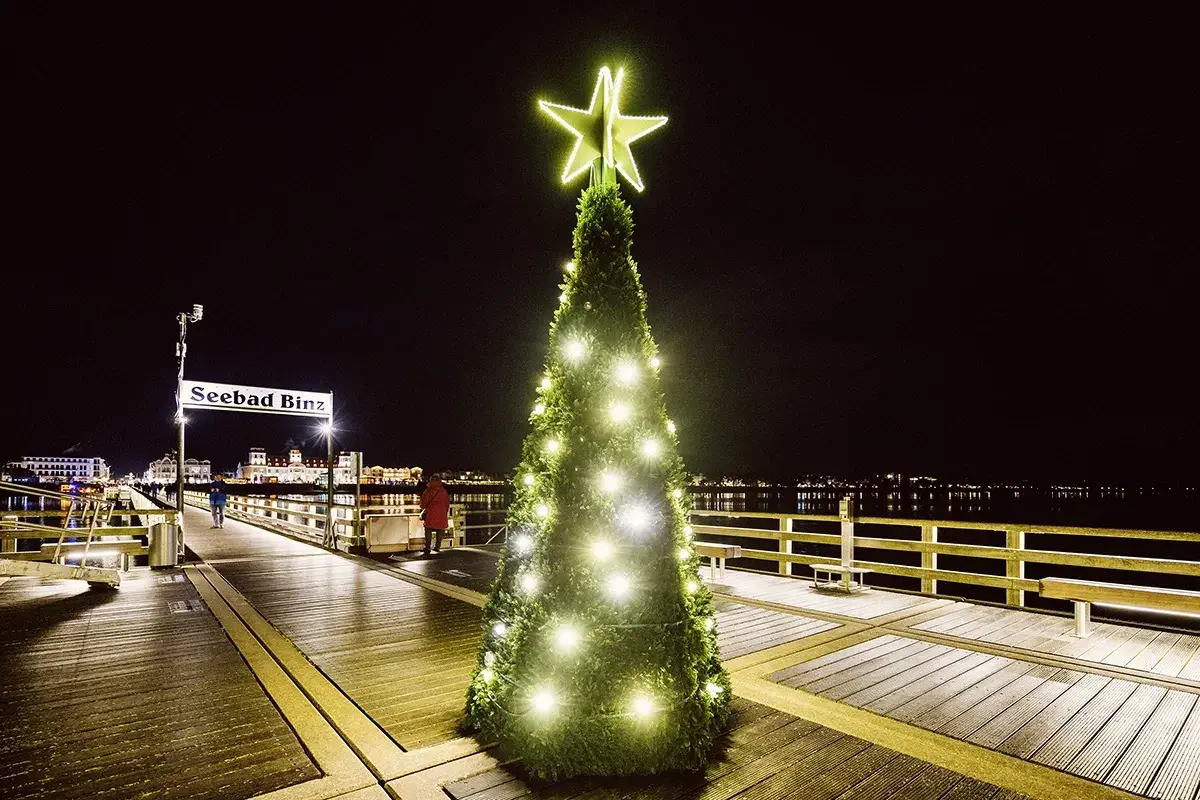 Ein festlich beleuchteter Weihnachtsbaum mit Sternspitze auf dem Holzsteg des Seebads Binz. Im Hintergrund die Lichter der Stadt.