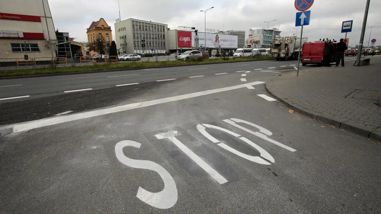 Na asfalcie widoczna jest biała linia warunkowego zatrzymania "STOP". W tle budynki i ruch uliczny.