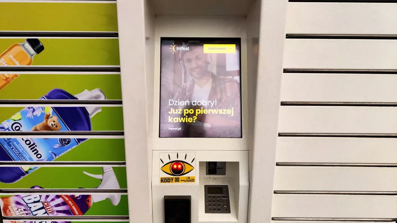 Ekran automatu InPost z uśmiechniętym mężczyzną i pytaniem "Dzień dobry! Już po pierwszej kawie?". Obok widać produkty z półek.