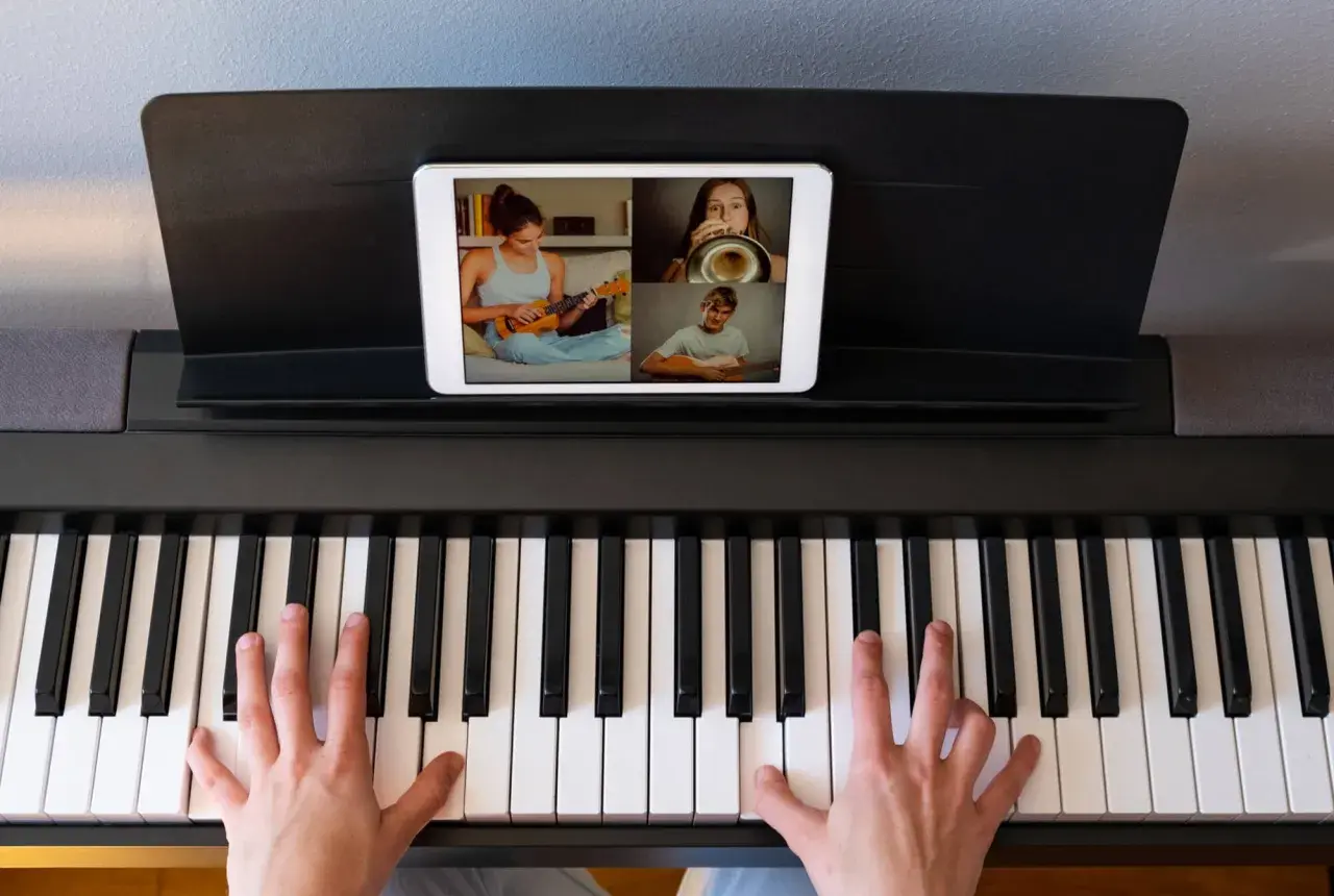 Ręce na klawiaturze pianina cyfrowego, na ekranie tabletu widać lekcję muzyki online z uczniami grającymi na ukulele, trąbce i gitarze.