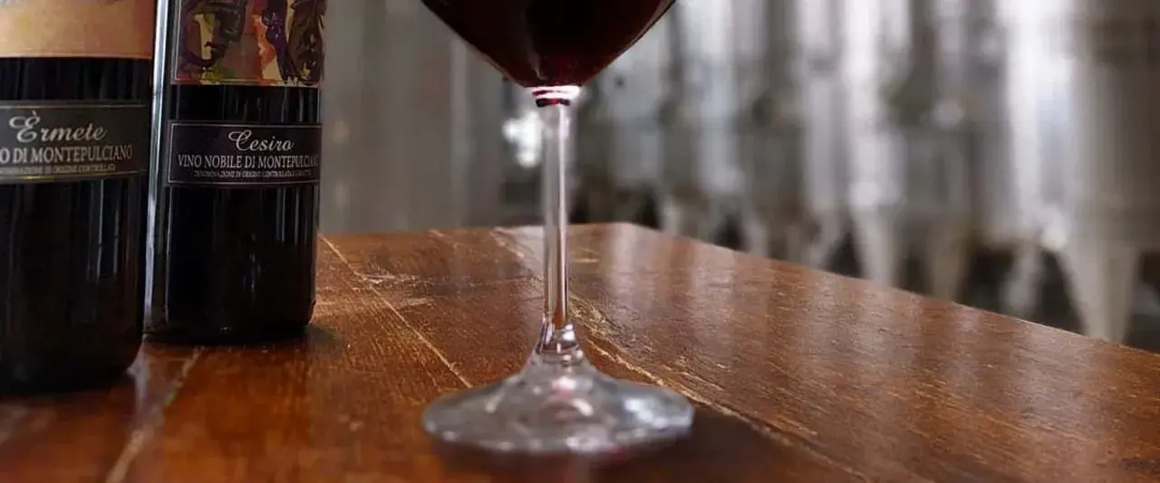 Bottiglie di Vino Nobile di Montepulciano e un calice di rosso, in un'ambientazione che evoca le cantine delle province di Siena.