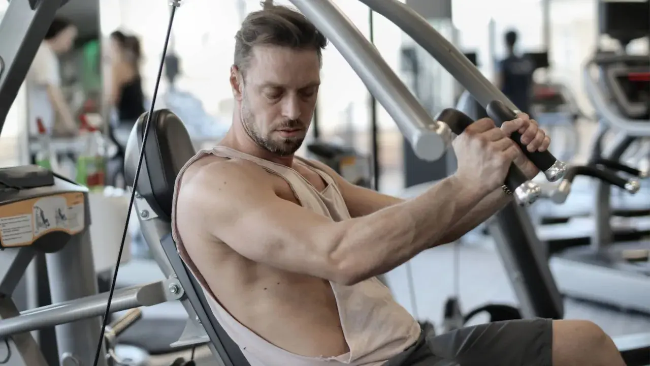 Mann trainiert im Fitnessstudio, um schnell zuzunehmen. Er sitzt auf einem Gerät und drückt die Griffe nach vorne.