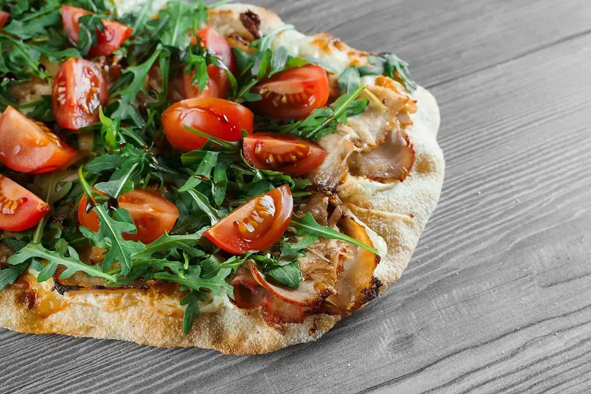 Une délicieuse pinsa recette garnie de tomates cerises, roquette fraîche et viande effilochée, sur fond de bois gris.
