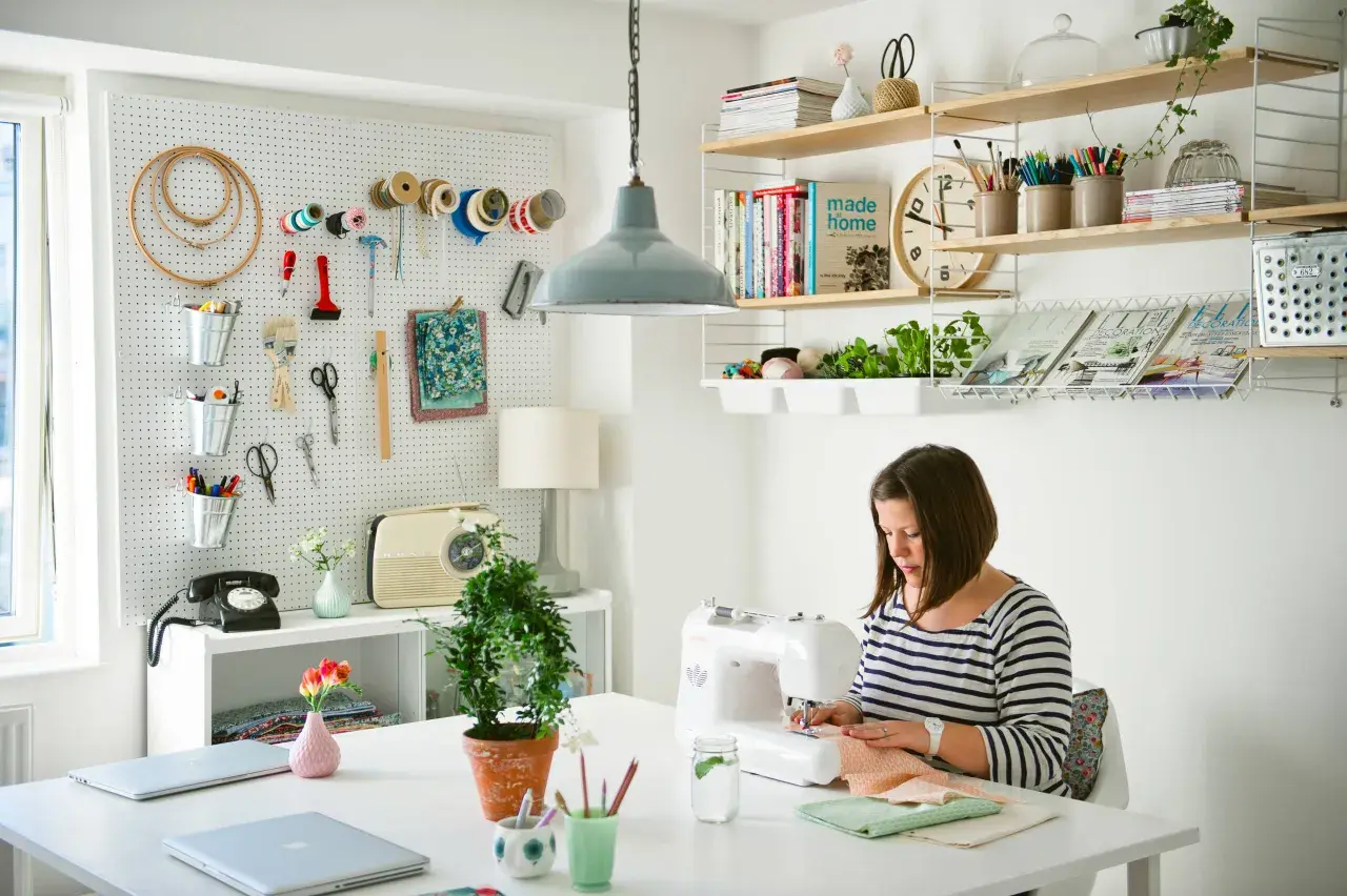 Mujer cosiendo en su **taller de manualidades en casa**. Pared con herramientas, estanterías con libros y plantas.