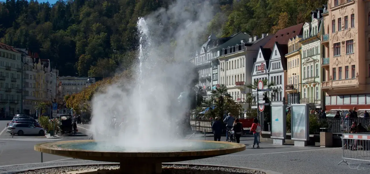 Der sprühende geysir karlsbad mit dampfender Fontäne vor bunten Häusern und grünen Bäumen.