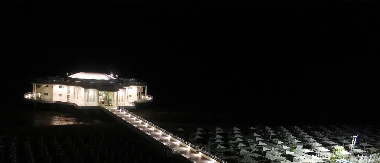 La Rotonda a Mare di Senigallia illuminata di notte, con la passerella che conduce all'edificio e la spiaggia con ombrelloni chiusi.