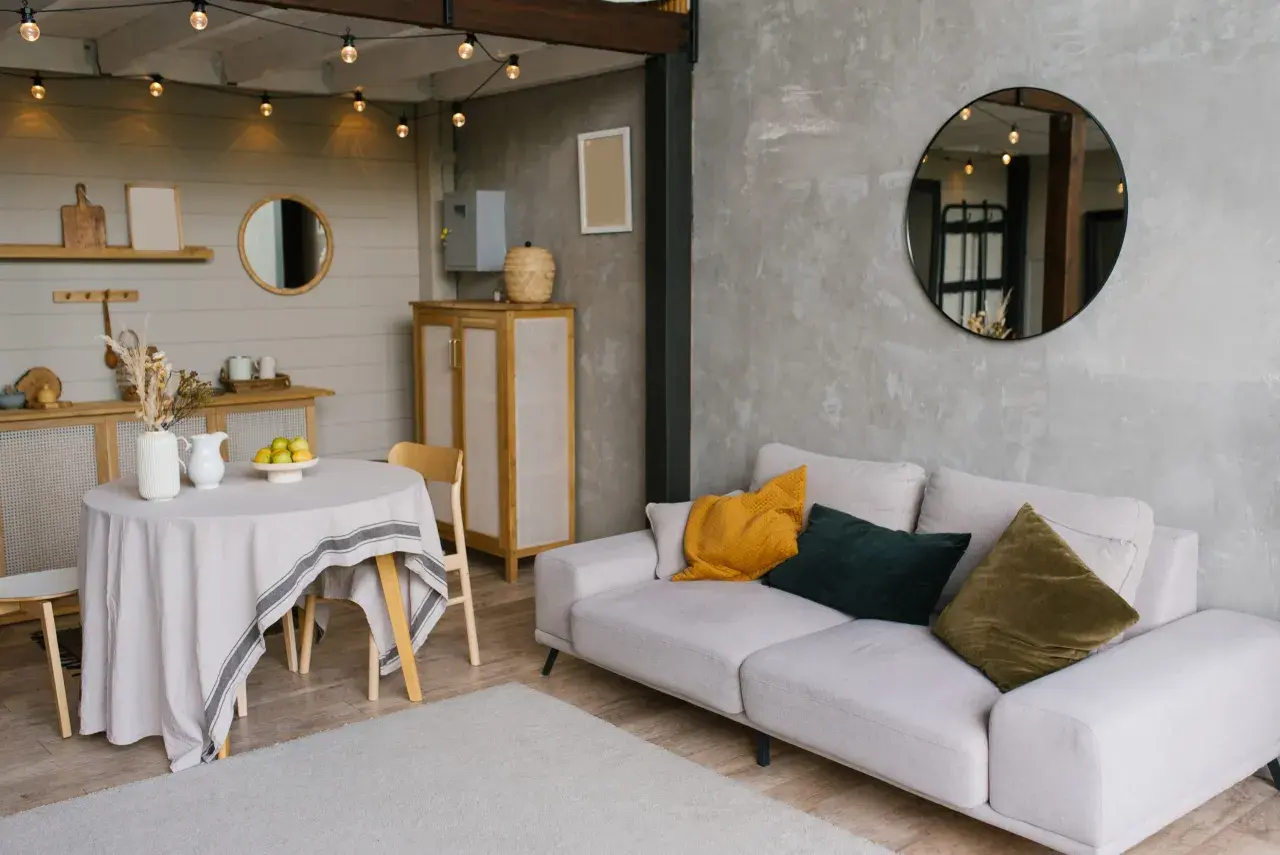 Un salon moderne avec un canapé clair, des coussins colorés et un miroir rond. La table ronde couverte d'une nappe grise et les guirlandes lumineuses ajoutent une touche chaleureuse, reflétant une nouvelle tendance déco.