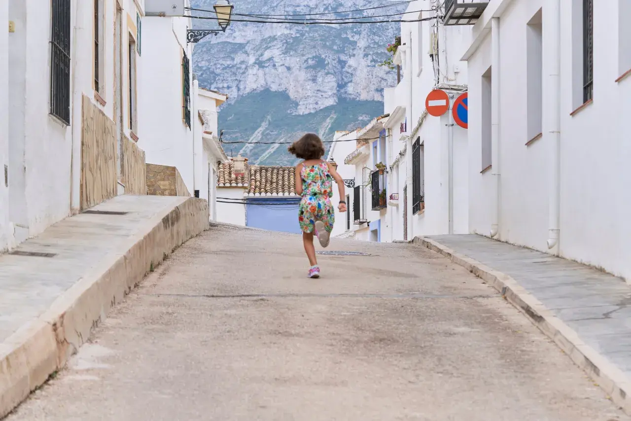 Niña corre por calle empinada de un pueblo de Alicante, con casas blancas y montañas al fondo.