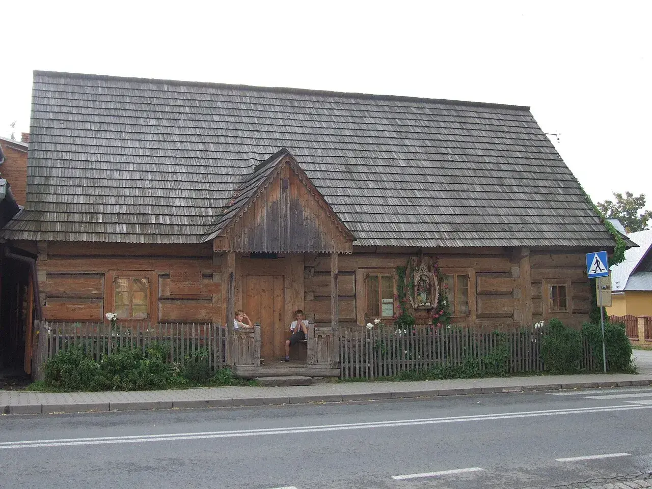 Drewniany budynek muzeum powstania chochołowskiego z gontowym dachem. Dzieci siedzą na progu.