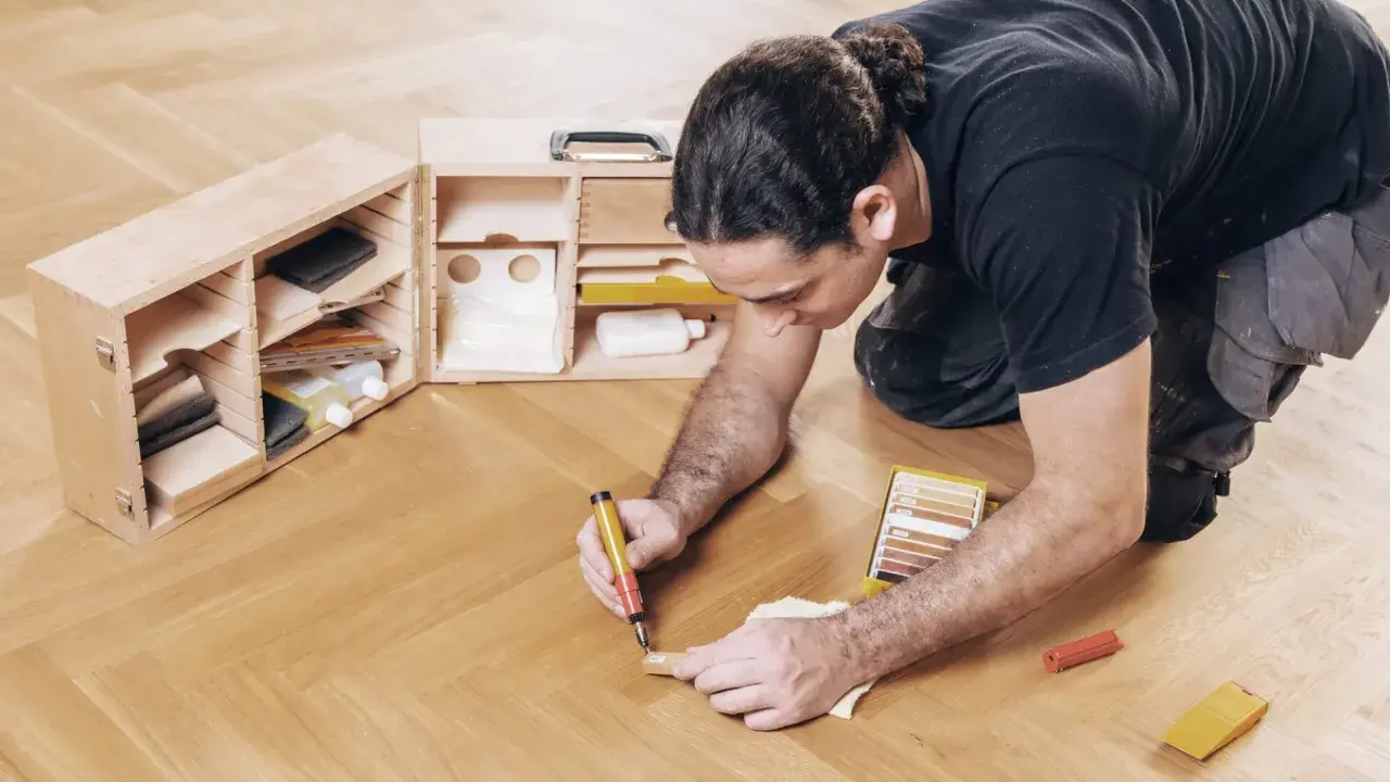 Mann repariert Parkett mit einem Reparaturstift und einer Farbkarte. Werkzeugkasten daneben.