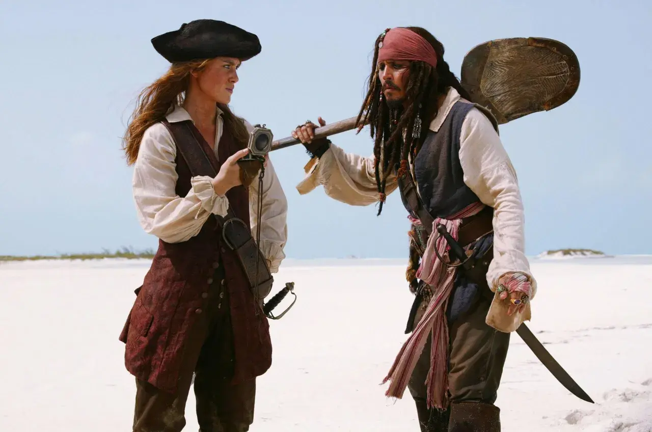 Elizabeth Swann i Kapitan Jack Sparrow, bohaterowie filmów "Piraci z Karaibów", stoją na plaży, rozmawiając.