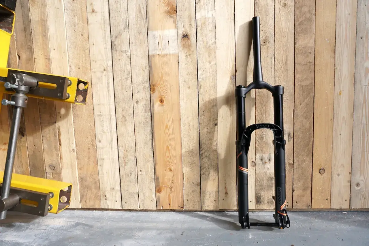 A black MRP Ribbon coil suspension fork stands against a wooden wall. The fork features orange and grey accents and the "ribbon" branding is visible.