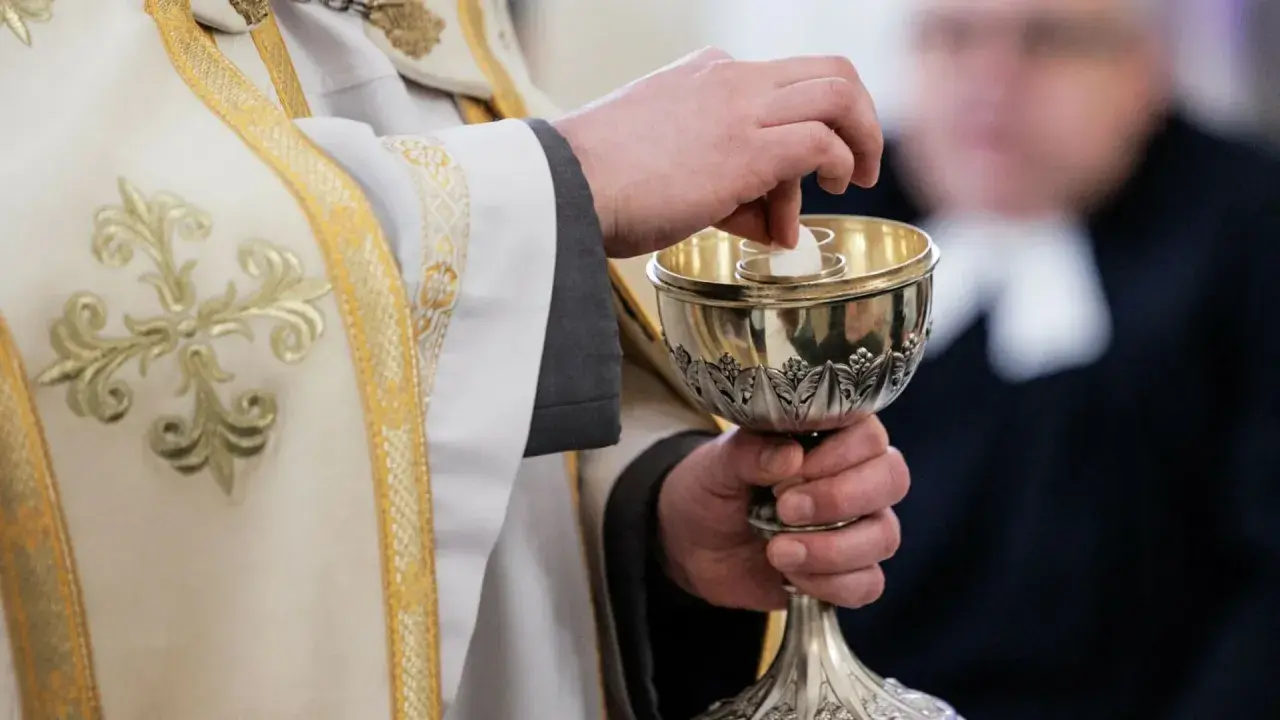 Dłoń księdza w białej szacie z haftem podaje hostię do kielicha. Komunia bez spowiedzi, symbol wiary.