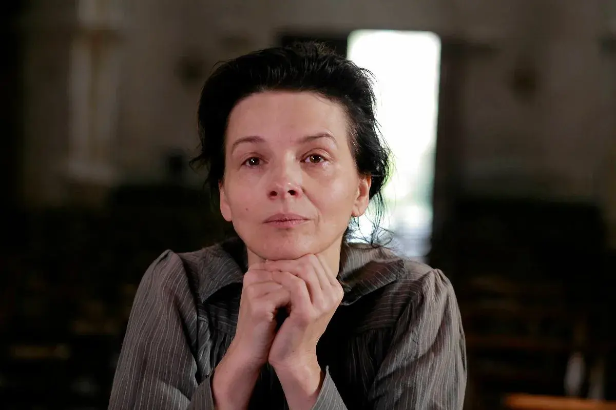 Portret kobiety w wieku dojrzałym, przypominającej Camille Claudel, z dłońmi podpartymi pod brodą, w stonowanym wnętrzu.