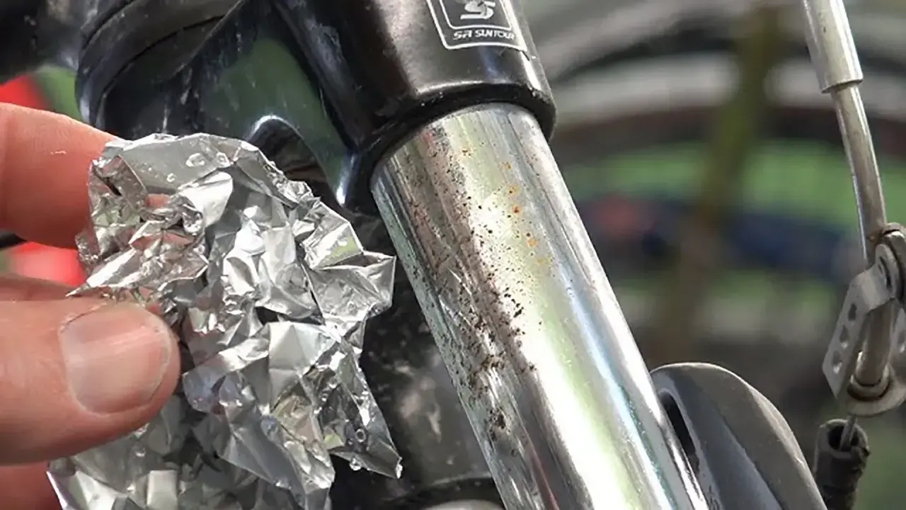 Mano che strofina con carta stagnola un ammortizzatore di bicicletta con macchie di ruggine e sporco.