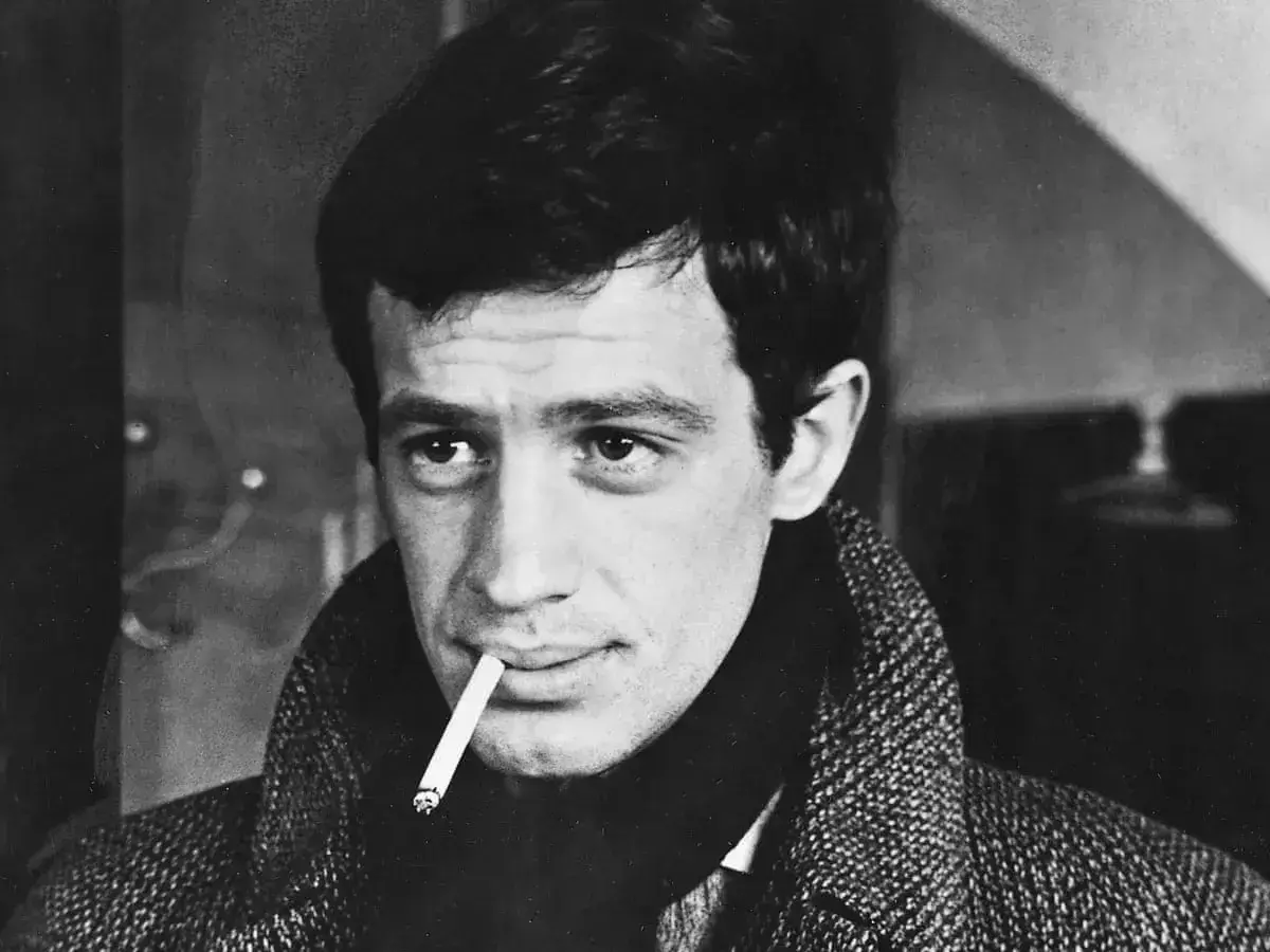 Jean-Paul Belmondo z papierosem w ustach, w płaszczu. Jego spojrzenie przywodzi na myśl niezapomniane **belmondo cytaty**.