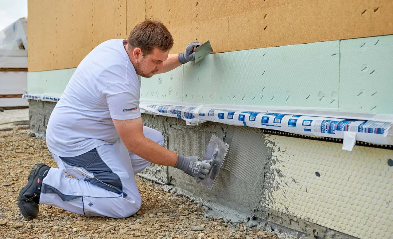Handwerker kniet und verputzt die Sockelzone einer Hausfassade mit einer Kelle.