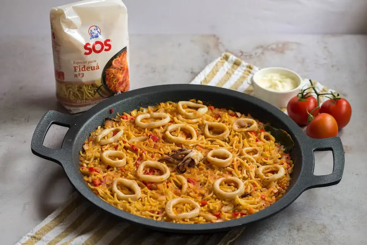 Fideuá con calamares lista para servir, acompañada de tomates y alioli.